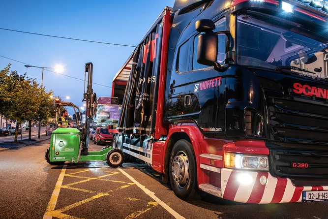 A forklift loads items onto a parked Scania G320 truck in an urban setting during dusk.