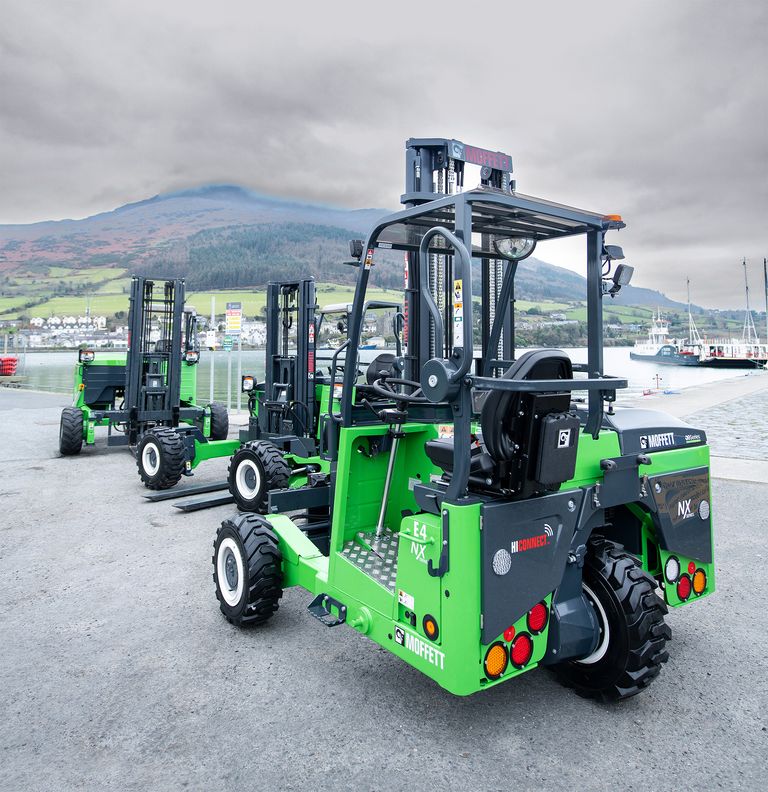 Three green forklifts are parked in a line on a concrete surface near a waterfront, with mountains visible in the background under a cloudy sky.