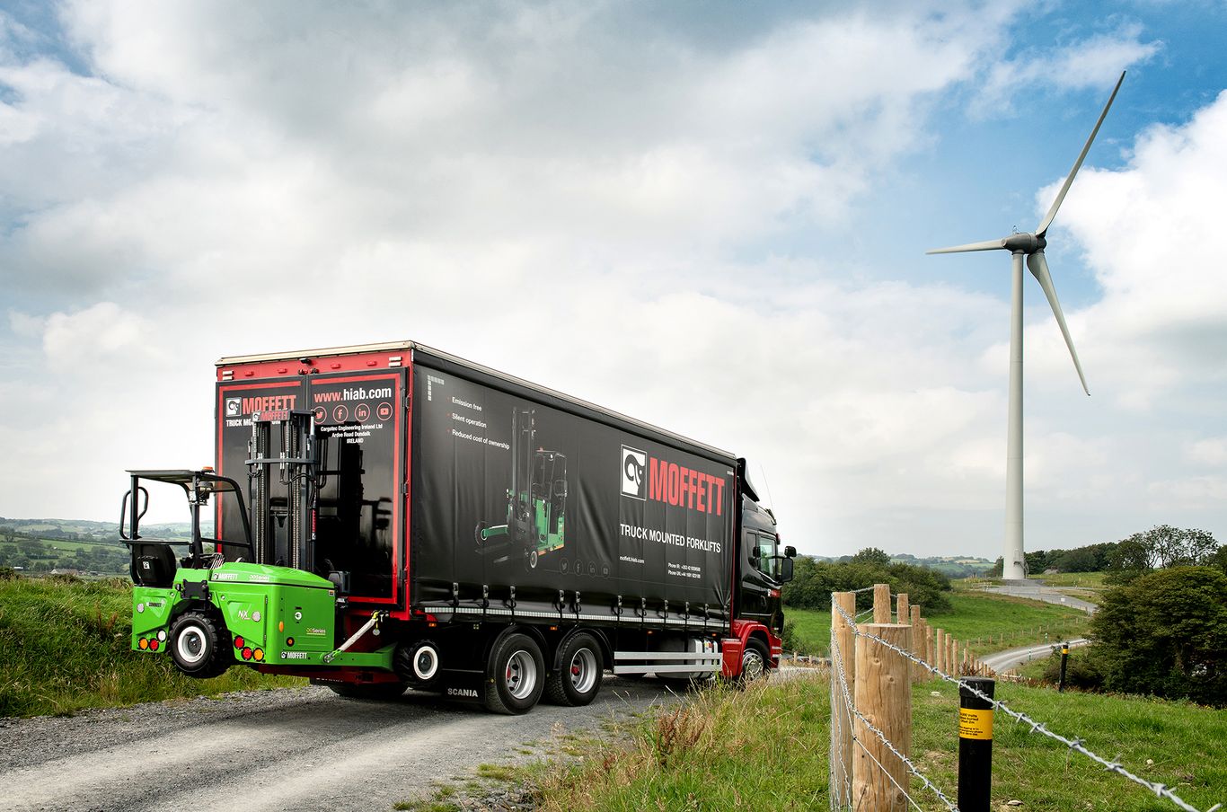 Truck with Moffett branding carrying a forklift drives on a rural road, with a wind turbine and green fields in the background.