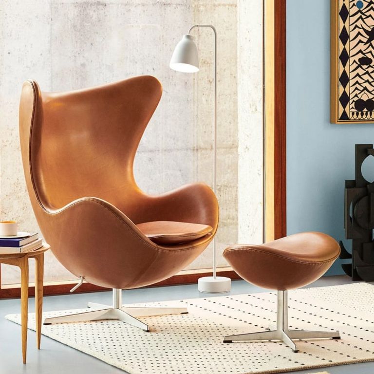 A modern living room featuring a brown egg chair with a matching ottoman, a small wooden side table, a white floor lamp, and a patterned wall hanging.