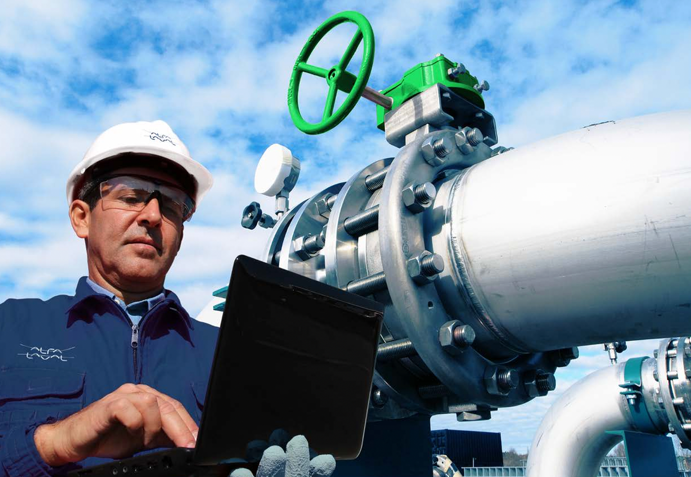 A worker in protective gear uses a laptop beside large industrial pipes with a green valve and a cloudy sky in the background.