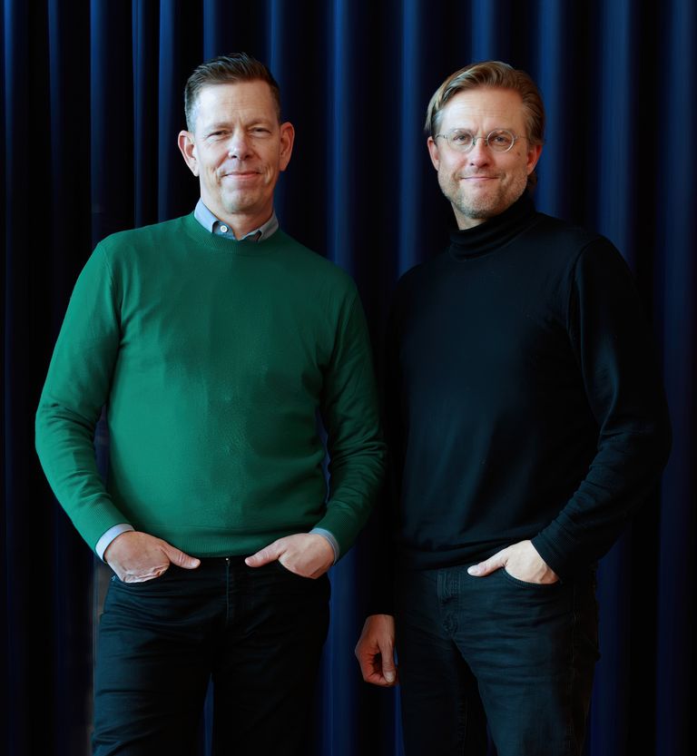 Two men stand side by side in front of a blue curtain. The man on the left wears a green sweater, while the man on the right wears a black turtleneck. Both have neutral expressions.