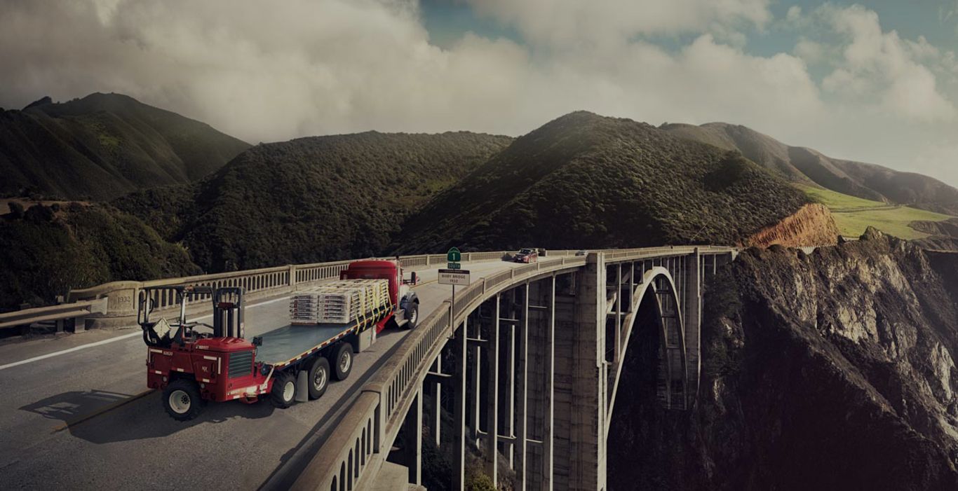 A flatbed truck carrying goods travels across a concrete arch bridge with mountainous terrain in the background.