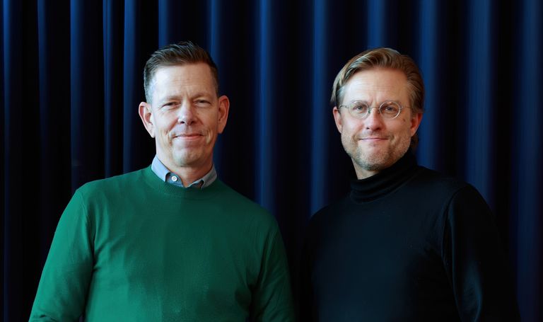 Two men standing in front of a dark blue curtain. One wears a green sweater, and the other wears a black turtleneck and glasses.